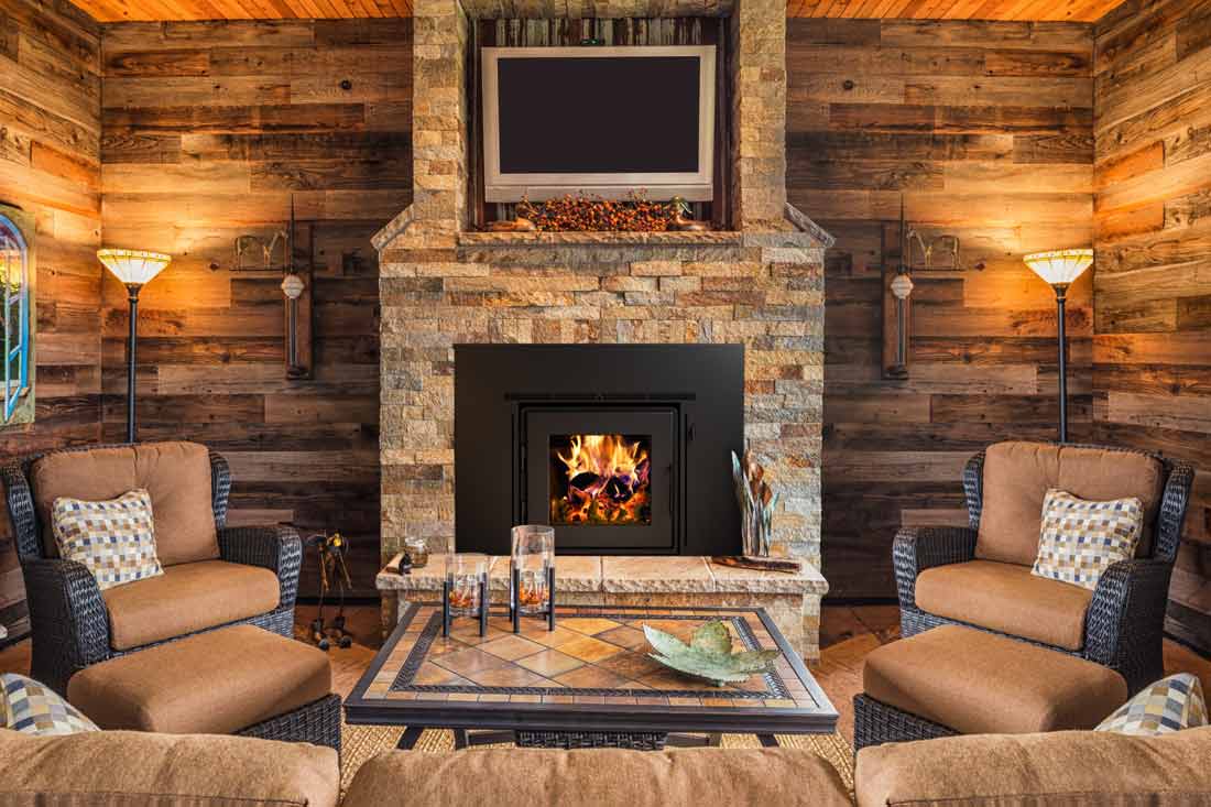 Stone fireplace with black fireplace insert and wood burning fire in room with barnwood walls and wicker furniture all in earthy tones