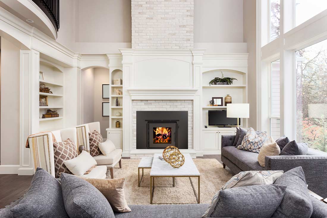 Brick fireplace with black fireplace insert and wood burning fire in room with white bookcase lined walls, tall windows, and upholstered furniture in cream and grey and neutral tones