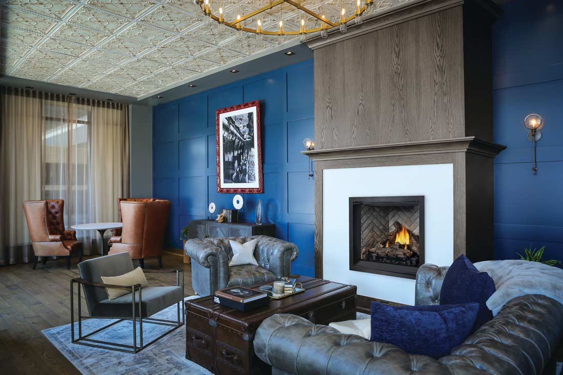 Sitting room with royal blue paneled wall along which is a Napoleon Elevation 36 gas fireplace with log set and yellow-orange flames. Fireplace Zen front is surrounded by white framed with wood mantel and cabinet.