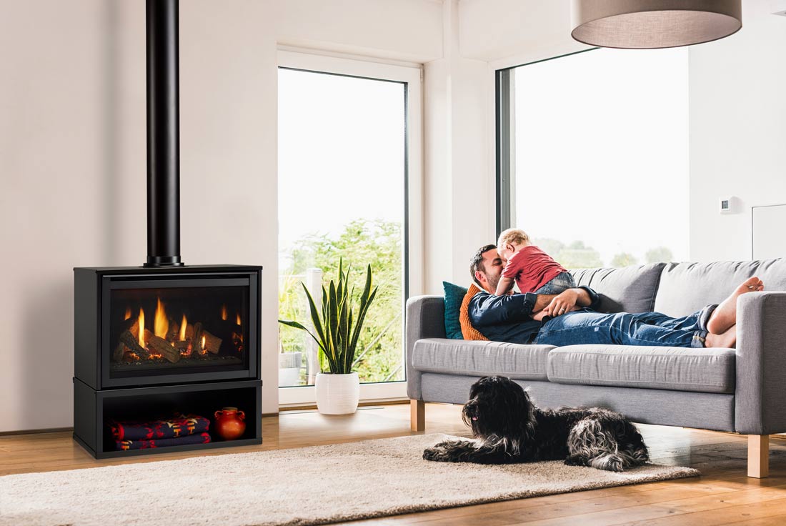 Free standing gas fireplace in cabinet with interior black glass panels reflecting flames throughout logs shown in white living room with wood floor, father and son on grey sofa with black dog nearby.