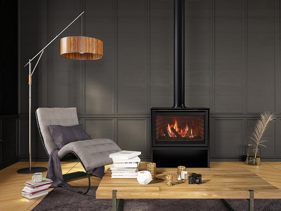 Free standing gas fireplace in cabinet with brick interior, and flames among logs shown in gray paneled sitting room with wood floor, containing a gray chair, floor lamp, and wood top table, book stacks, and coffee cup.
