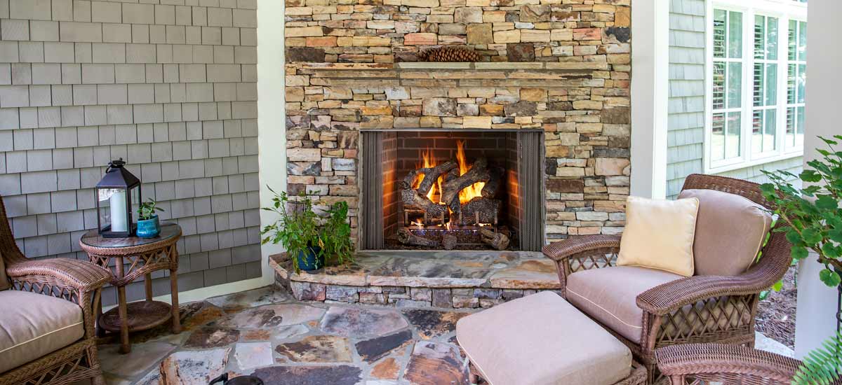 Gas logs burning in ledge stone outdoor fireplace along shingled exterior house walls flanked by wicker furniture