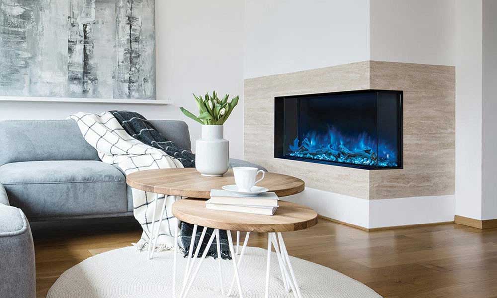 Landscape Pro Multi electric fireplace with right corner installation shown with blue flames and ember bed in living room wall, wood top tables, grey sofa with white and grey throws draped across.