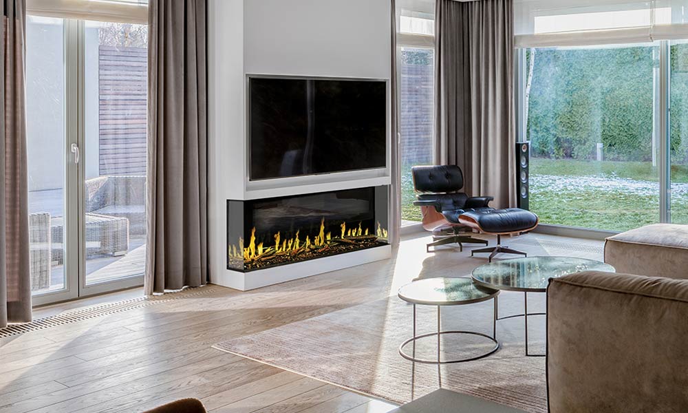 Three-sided linear electric fireplace with yellow flames on white wall under television flanked by floor to ceiling windows in living room with wood floor and black reclingin chair to the side.