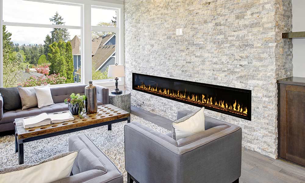 Very long linear electric fireplace with yellow flames on white rock wall in living room with grey furniture and picture window.
