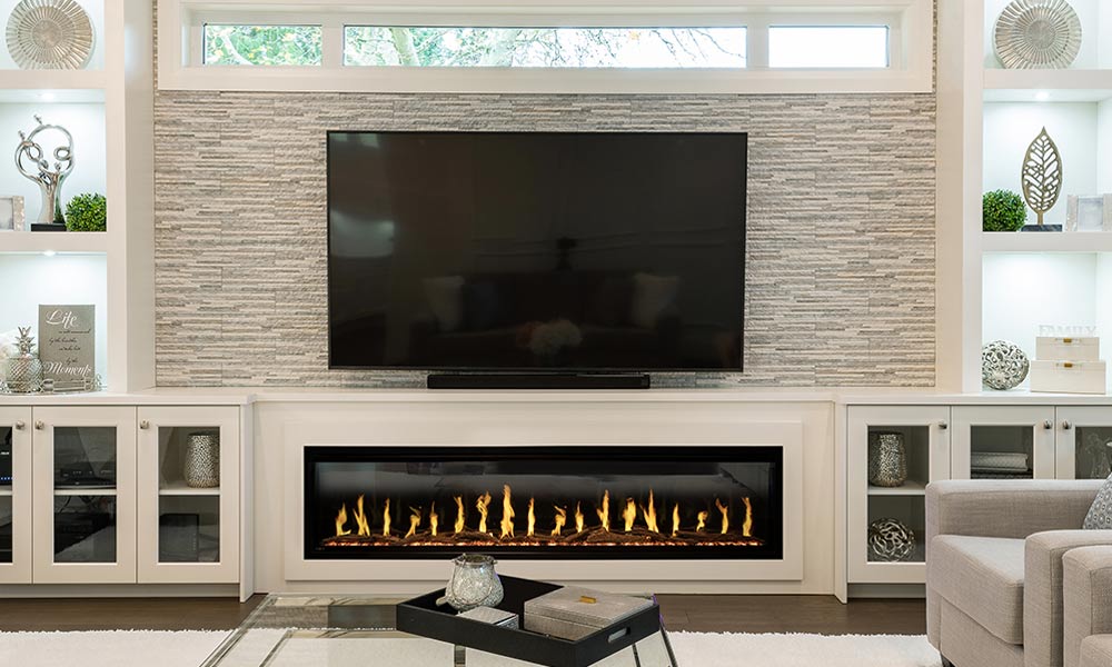 Long electric fireplace burning under television on white stone wall flanked by shelves with decor in living room.