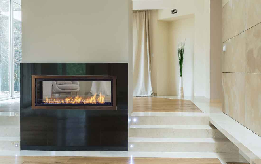 Monessen Artisan see-through linear vent free gas fireplace with copper front, black stone surround on short wall open on both sides and marble steps to upper level