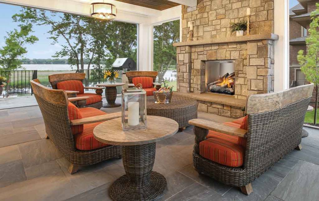 Stainless steel outdoor gas fireplace installed in stone on covered patio with orange cushioned rattan furniture and tables, lake view.