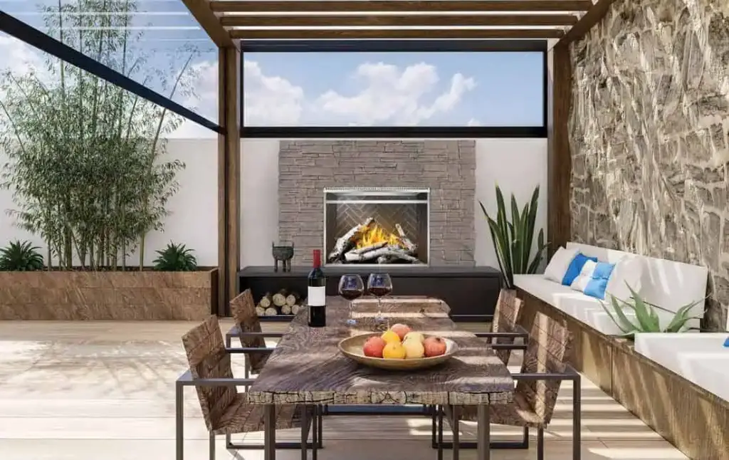 Outdoor patio with bench seats along stone wall, Riverside gas fireplace at end, wood and metal table and chairs with wine and fruit